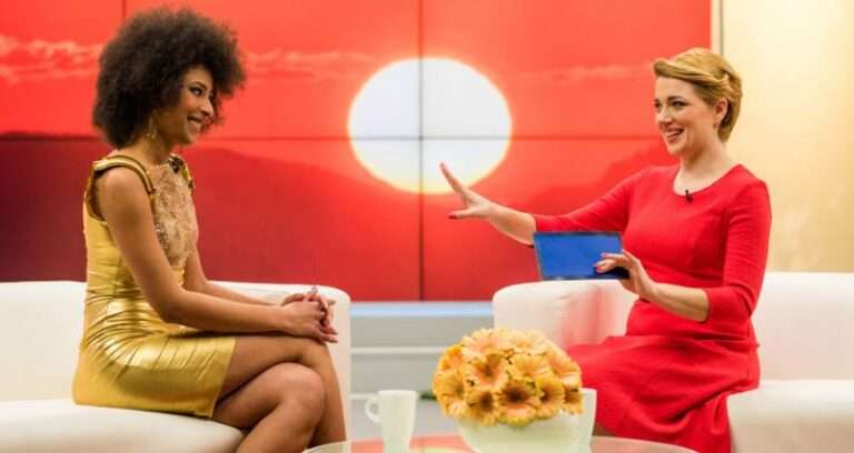 A journalist in a red dress interviews a guest in a gold dress on a brightly lit TV set with a large sun backdrop. Both women are smiling and engaged, capturing the essence of how journalists and TV hosts make people feel seen by giving their full attention.