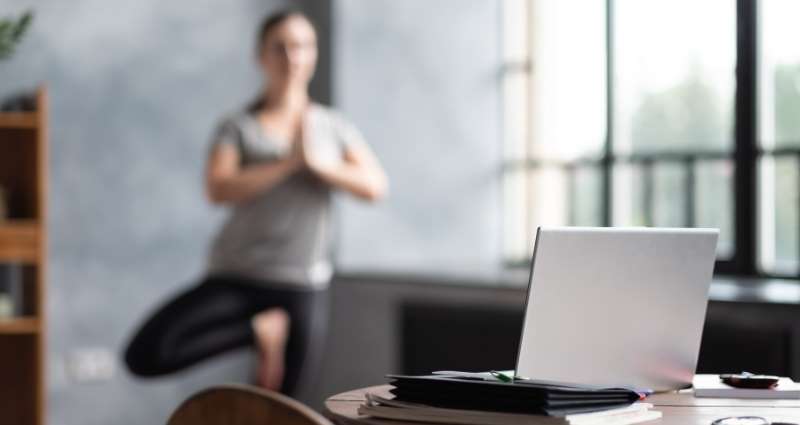 A woman practicing her daily rituals by doing yoga in a tree pose near a table with a laptop and notebooks, in a bright, modern home setting.