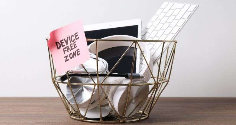 A wire basket labeled "Device Free Zone" filled with electronics like headphones, a tablet, and a keyboard, symbolizing the daily ritual of unplugging from technology.