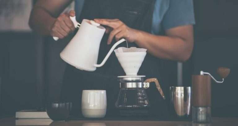A person performing their daily rituals by brewing pour-over coffee using a white kettle and a ceramic dripper, surrounded by a mug, coffee grinder, and metal cup on a wooden counter.