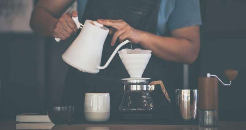 A person performing their daily rituals by brewing pour-over coffee using a white kettle and a ceramic dripper, surrounded by a mug, coffee grinder, and metal cup on a wooden counter.