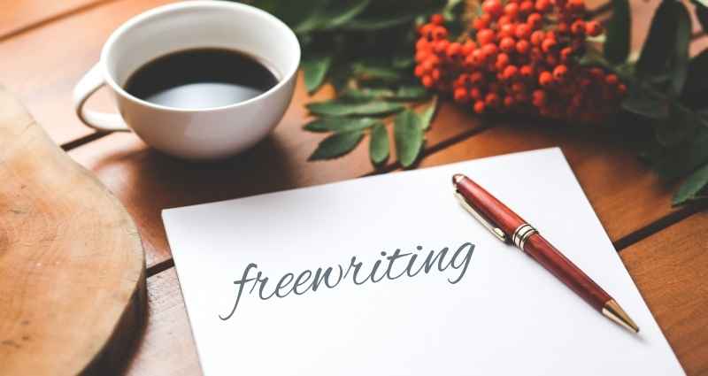 A cup of coffee sits beside a sheet of paper with the word “freewriting” written in cursive, accompanied by a red pen and sprigs of greenery and berries. This image reflects creative methods, such as freewriting, which encourages spontaneous and unfiltered expression to unlock new ideas and overcome creative blocks.