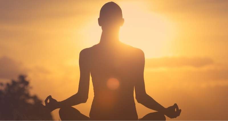 A person sits in a meditative pose at sunrise, silhouetted against a glowing sky. The peaceful setting reflects creative methods, such as meditation, which help enhance creative thinking.