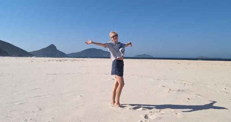 A person joyfully stands barefoot on a vast, sunny beach with arms outstretched, surrounded by mountains and clear blue sky. This image reflects creative methods, such as taking a vacation, which offers mental rest and new perspectives.